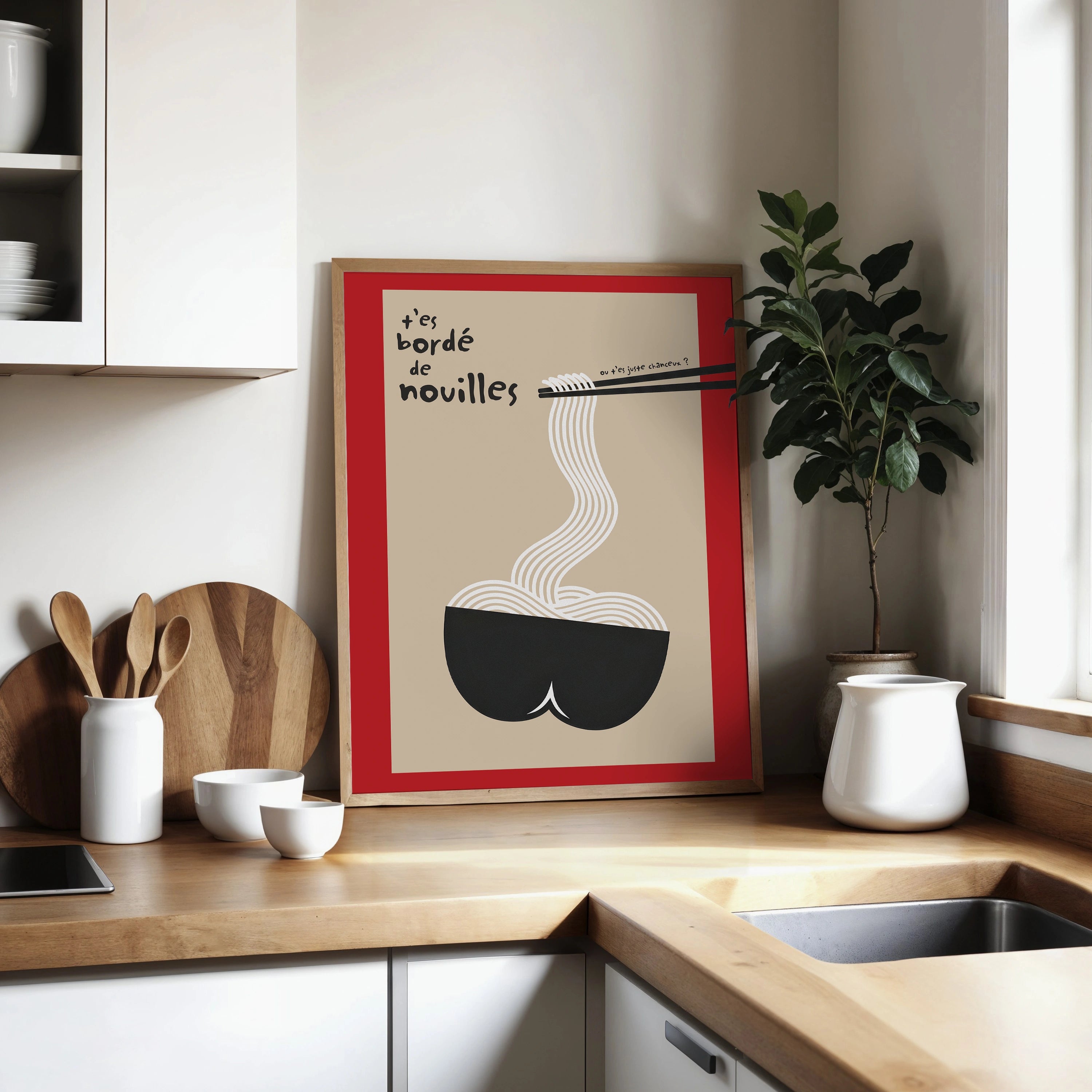 Affiche humour cuisine posée sur un plan de travail bois avec planches à découper et plante verte. Cadre bois, fond sable, bordure rouge, bol noir stylisé rappelant des fesses, long filet de nouilles blanches vers des baguettes, lettrage noir « t’es bordé de nouilles » + aparté manuscrit. Décoration murale moderne pour coin café, cuisine chaleureuse, style scandinave japandi.