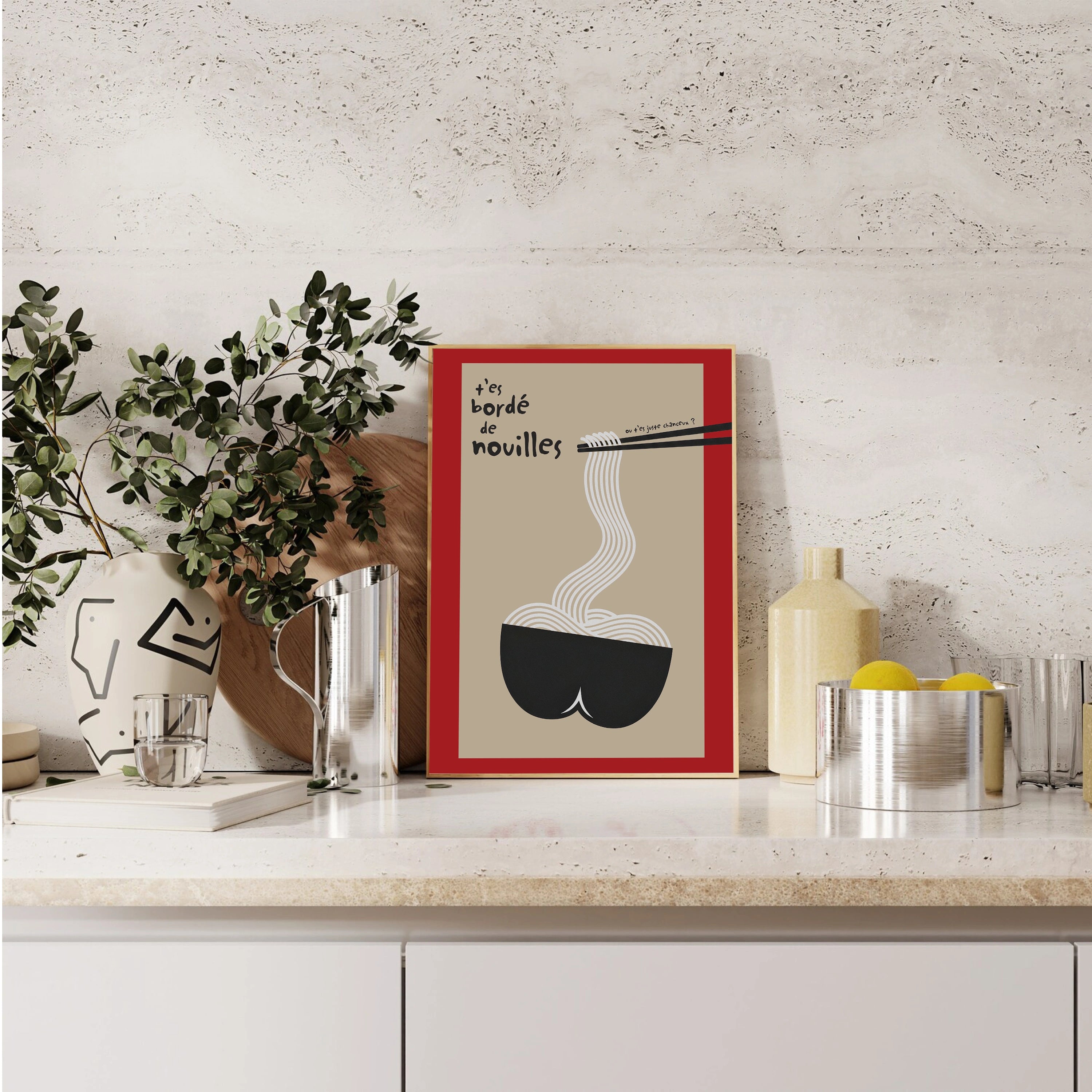 Poster déco culinaire dans une cuisine contemporaine : crédence minérale claire, objets inox et céramique. Affiche au graphisme minimal : fond beige, cadre rouge interne, bol noir aux formes arrondies évoquant des fesses, nouilles blanches fines, baguettes noires, typographie manuscrite « t’es bordé de nouilles ». Idéal cadeau humour, déco design cuisine, esprit graphique.