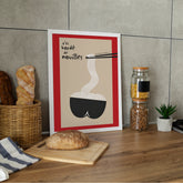 Affiche encadrée blanche sur plan de travail bois, baguettes, pain et bocaux de pâtes en arrière-plan. Illustrations : fond sable, large bordure rouge, bol noir forme fessier, nouilles blanches ondulées vers baguettes noires, texte manuscrit « t’es bordé de nouilles ». Style minimal, clin d’œil culinaire, parfait pour coin cuisine, bistrot maison, déco drôle.