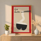 Affiche encadrée or posée sur enfilade bois, lumière dorée, vase blanc et herbes de pampa. Design minimal humoristique : fond beige, cadre rouge interne, bol noir en forme de fesses, nouilles blanches attrapées par baguettes noires, texte manuscrit « t’es bordé de nouilles ». Décoration murale cuisine / salle à manger, style moderne, clin d’œil pop.