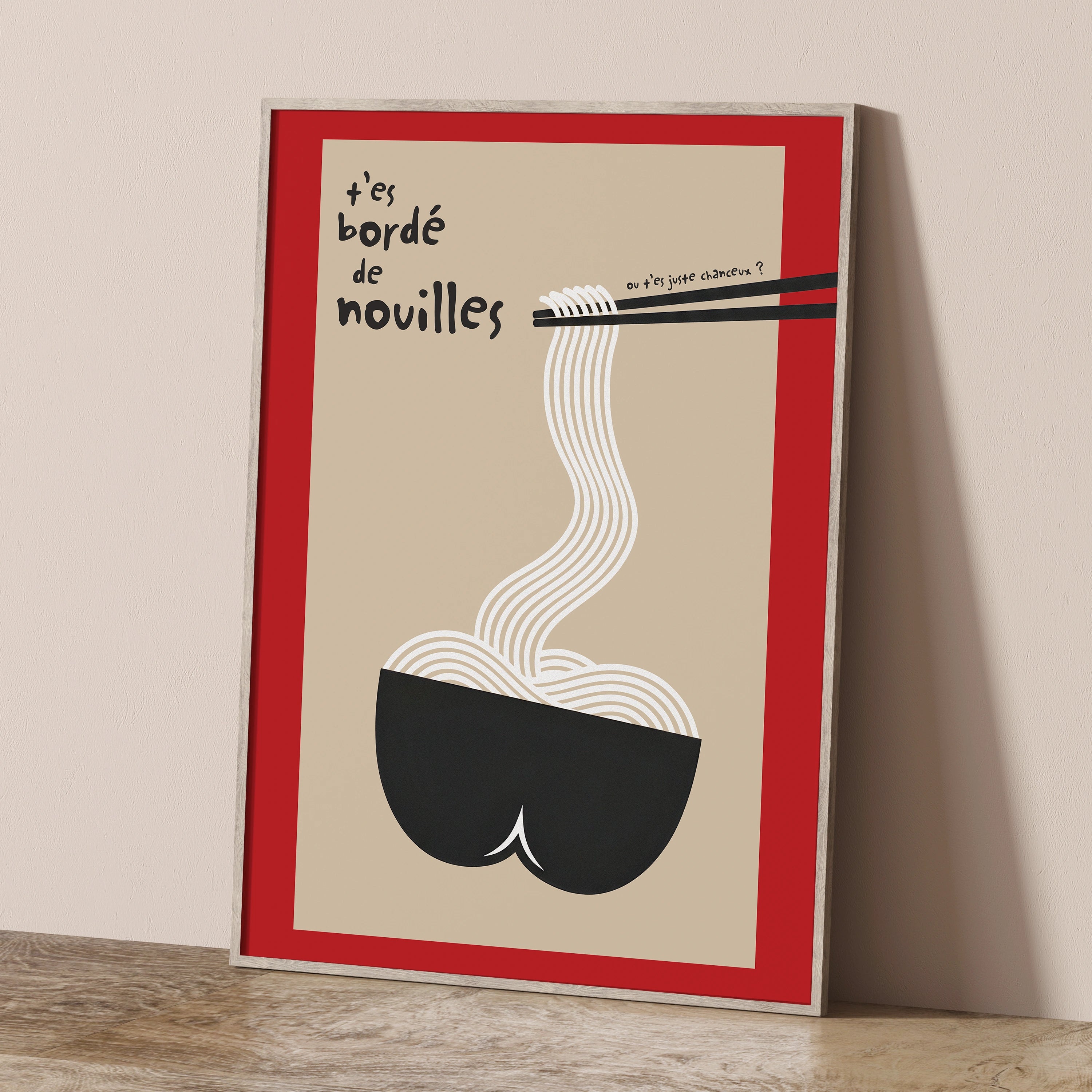Affiche « t’es bordé de nouilles » posée contre un mur beige, cadre bois clair, fond sable encadré d’un liseré rouge. Illustration minimaliste humoristique : un bol noir stylisé en forme de fesses d’où s’élèvent des nouilles blanches attrapées par des baguettes noires, typo manuscrite noire, petite phrase « ou t’es juste chanceux ? ». Idéal déco cuisine, coin café, style japandi, pop, jeu de mots français.