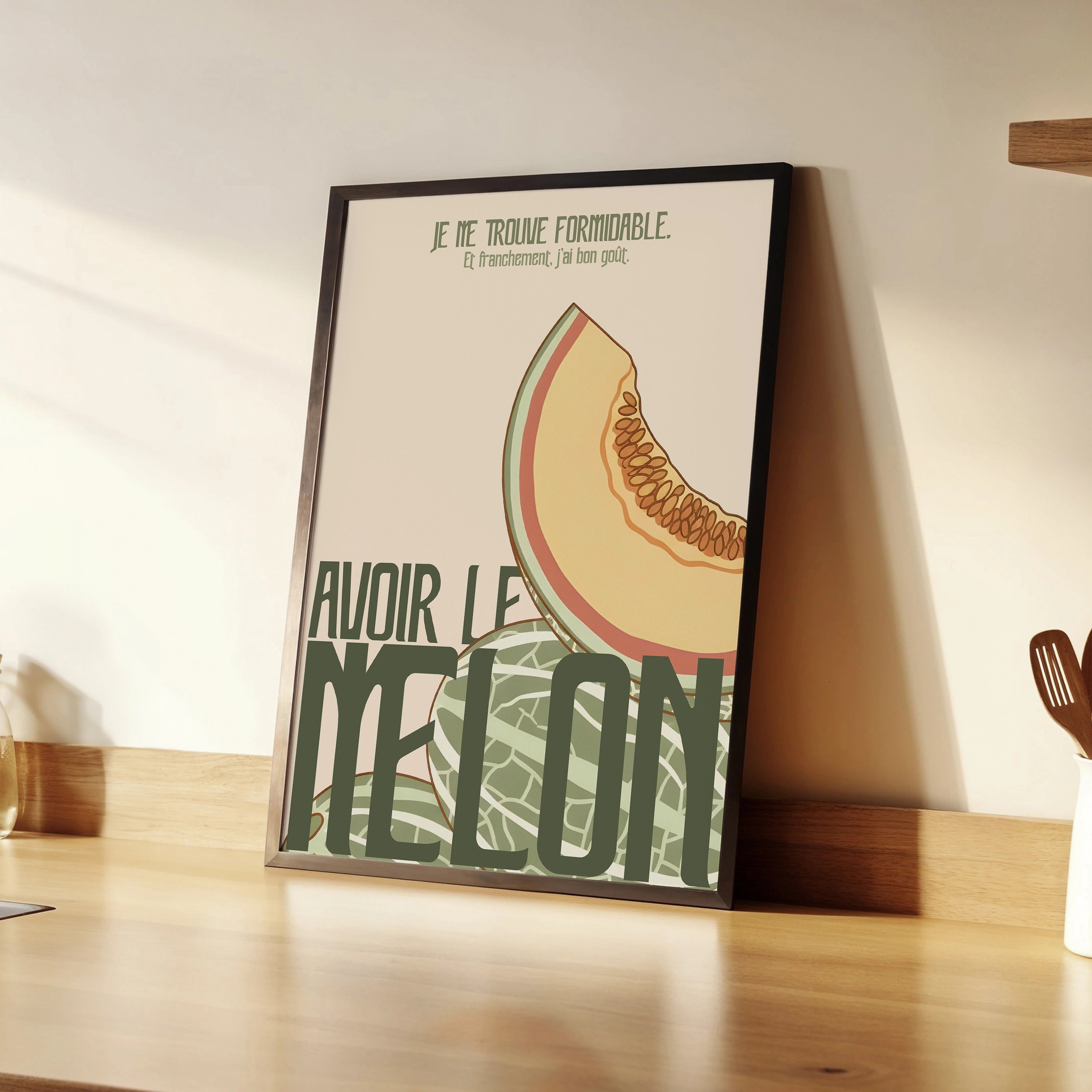 Affiche « Avoir le melon » inclinée sur un plan de travail, cadre noir fin. Illustration flat en couleurs pastel : demi-melon orange et jaune, écorce verte texturée, typographie massive vert foncé. Lumière chaude, ambiance cuisine contemporaine avec ustensiles en bois et plan de travail clair.