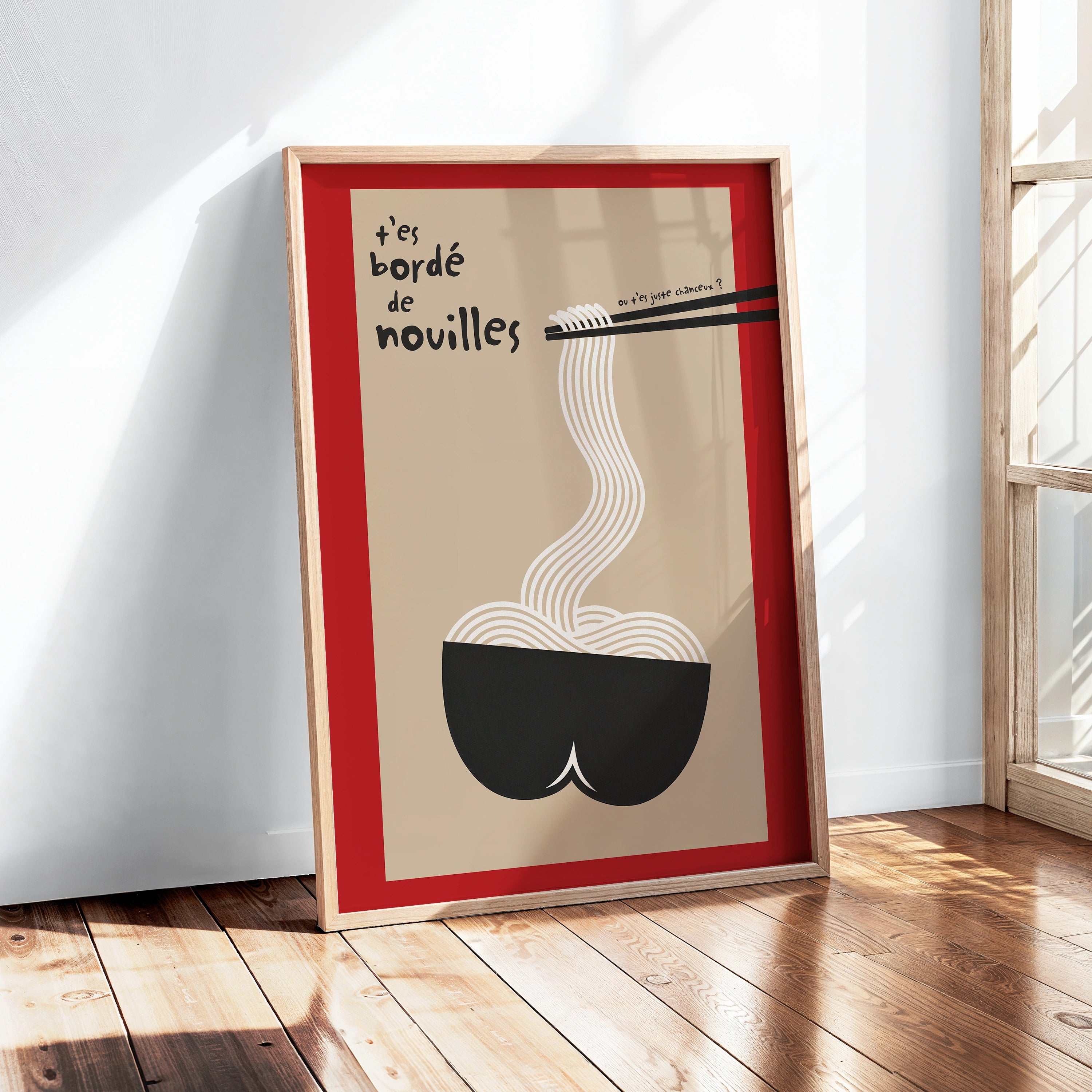 Poster encadré près d’une fenêtre, lumière naturelle et reflets sur le verre. Design : fond beige, large bordure rouge, bol noir en forme de popotin, nouilles blanches aux lignes courbes saisies par des baguettes, texte manuscrit « t’es bordé de nouilles ». Ambiance lumineuse, parquet, cadre bois clair. Affiche déco cuisine / salle à manger, style minimal, second degré, inspiration japonaise.