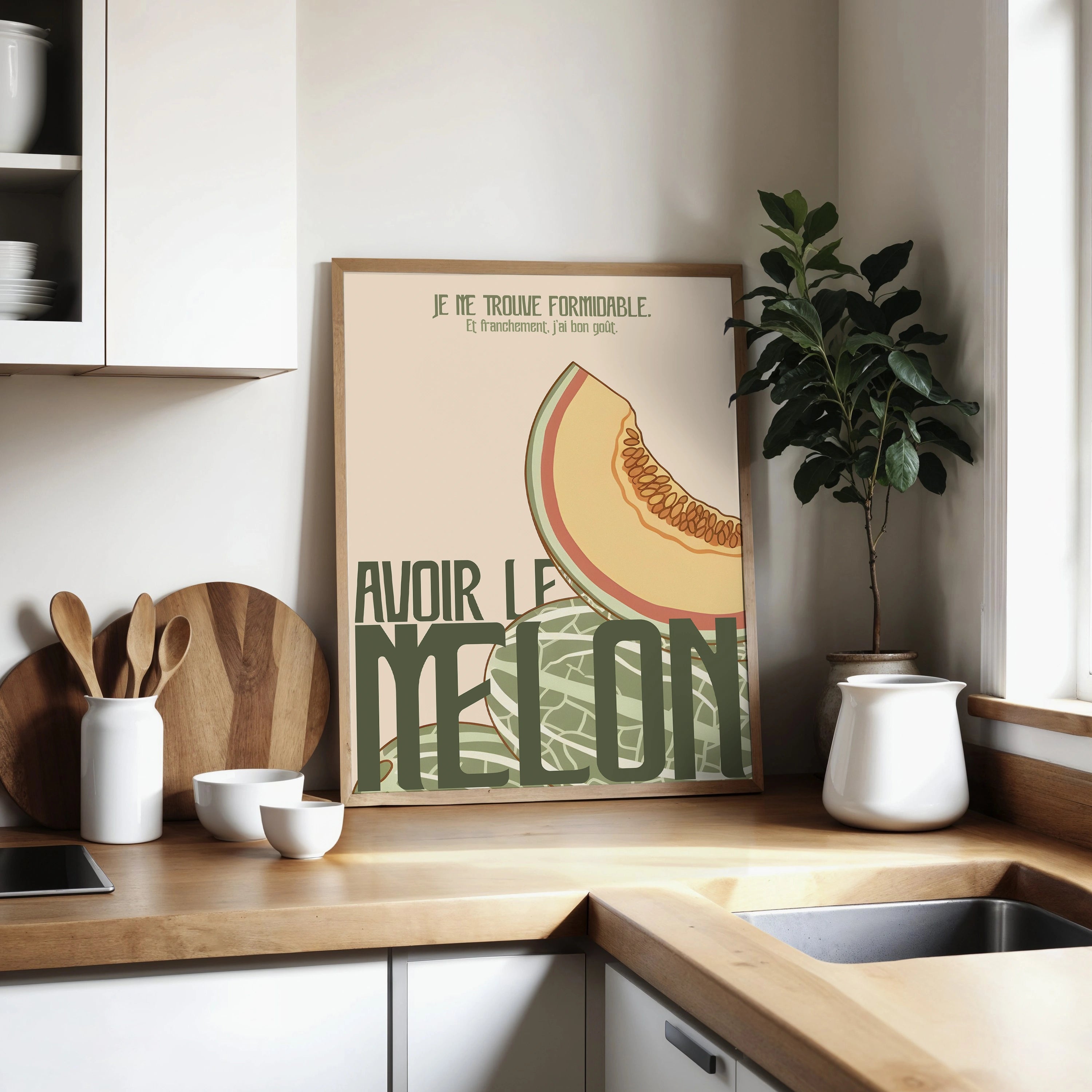 Affiche « Avoir le melon » posée dans un coin de cuisine lumineuse, cadre bois clair. Illustration de melon en style minimaliste pastel, texte décalé en vert foncé. Décor scandinave : plan de travail bois, pot en céramique blanc, plante verte, fenêtre apportant une lumière douce.