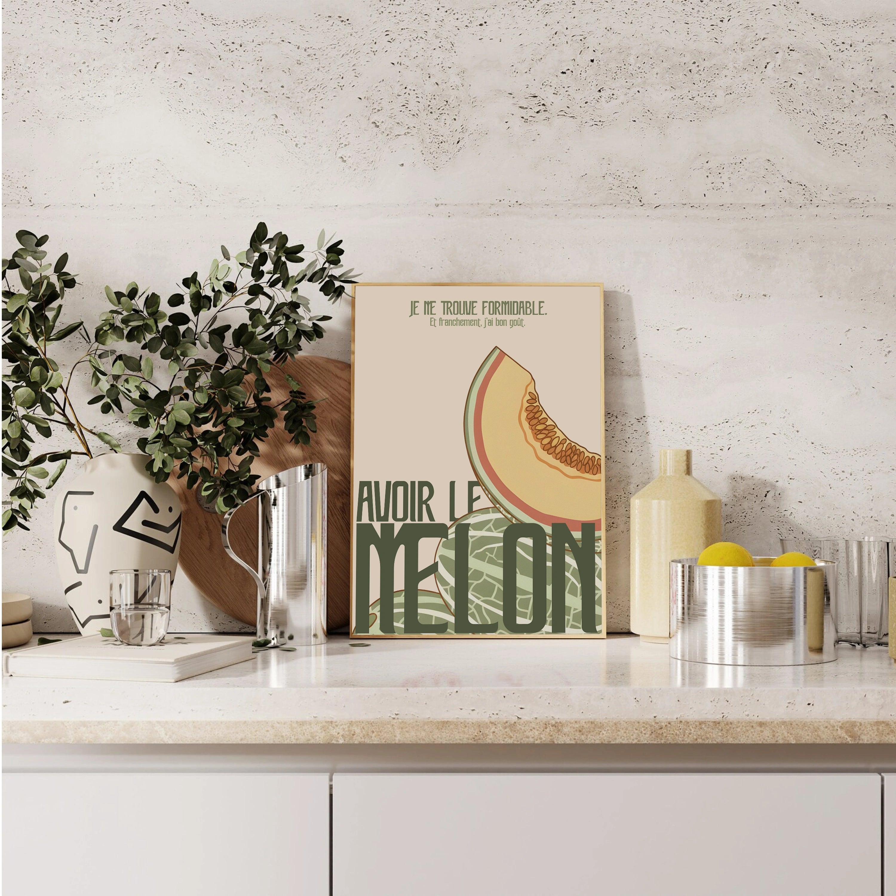 Poster « Avoir le melon » placé sur un plan de travail moderne, devant un mur clair texturé. Illustration flat pastel : tranche de melon orange, peau vert sauge, typographie audacieuse vert foncé. Mise en scène déco avec carafe métal, planche ronde, plante verte et bols citronnés, ambiance cuisine design.