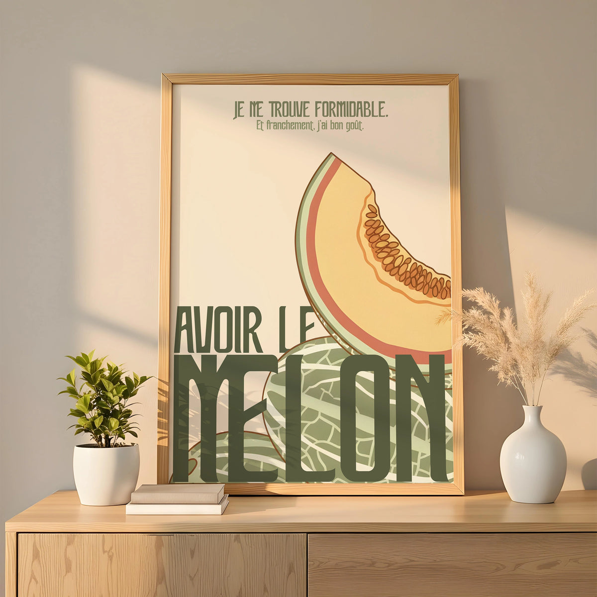 Affiche humoristique « Avoir le melon » posée sur un meuble en bois clair, encadrée d’un cadre en bois naturel. Illustration flat : demi-melon dessiné en tons pastel (jaune, vert sauge, orange), motif d’écorce texturé, typographie large vert foncé. Lumière douce, déco minimaliste avec plantes et vase blanc dans un intérieur moderne.