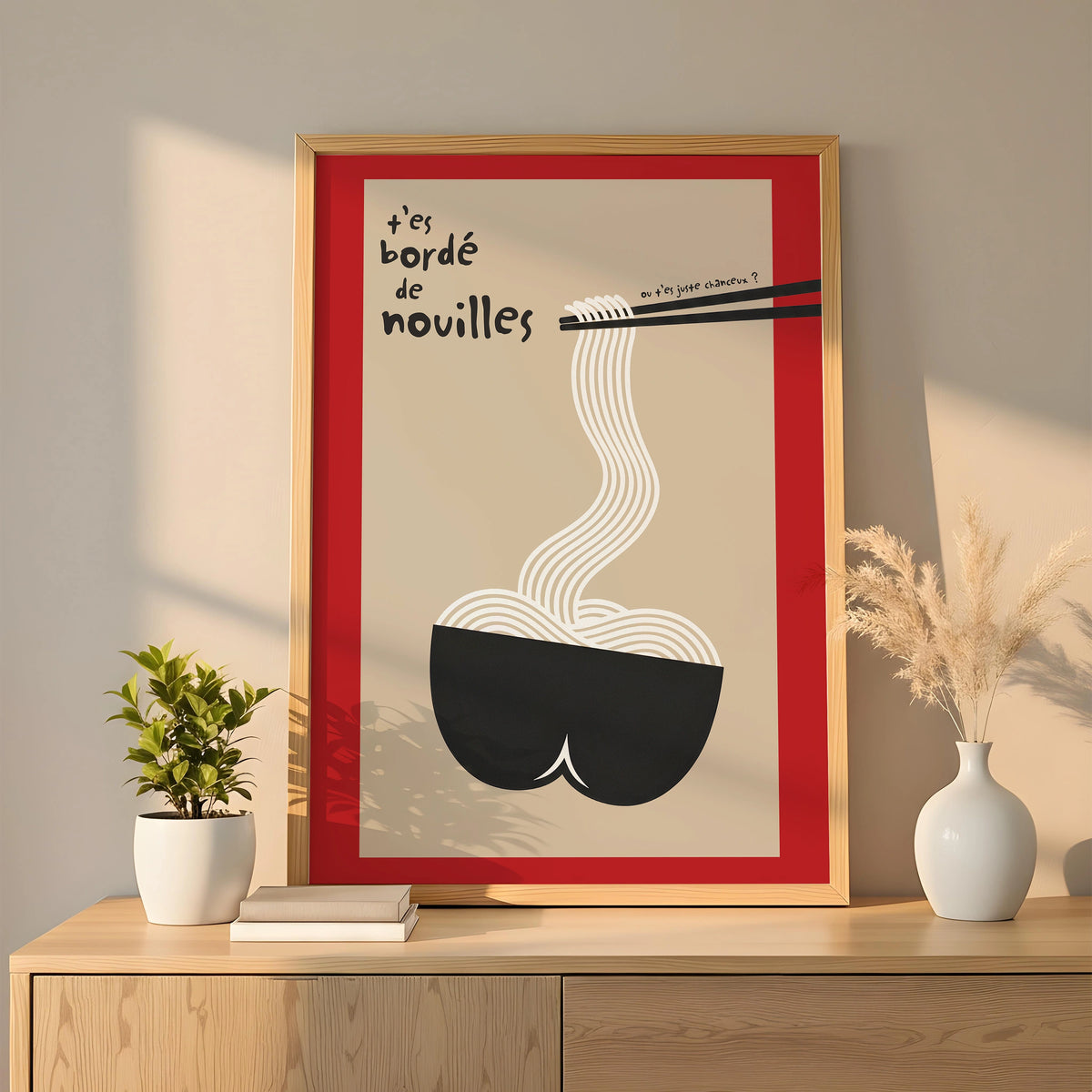 Affiche encadrée or posée sur enfilade bois, lumière dorée, vase blanc et herbes de pampa. Design minimal humoristique : fond beige, cadre rouge interne, bol noir en forme de fesses, nouilles blanches attrapées par baguettes noires, texte manuscrit « t’es bordé de nouilles ». Décoration murale cuisine / salle à manger, style moderne, clin d’œil pop.