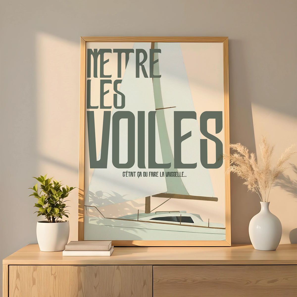 Affiche humoristique “Mettre les voiles – C’était ça ou faire la vaisselle” posée dans un cadre bois clair sur un buffet minimaliste. Illustration d’un voilier en flat design avec grandes voiles vert d’eau et beige, lignes épurées, typo rétro étirée. Lumière douce, déco naturelle avec plante verte et vase pampas.